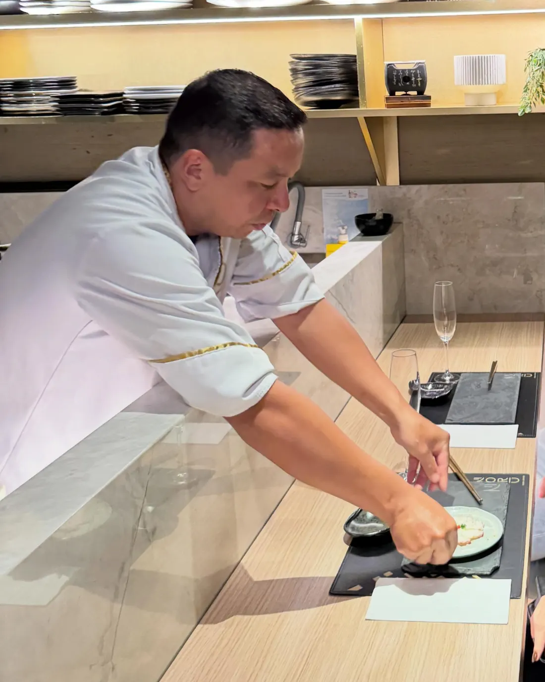 Um chef de dólmã branco com friso dourado serve uma peça no balcão de madeira, sob as prateleiras iluminadas com louças e plantas.