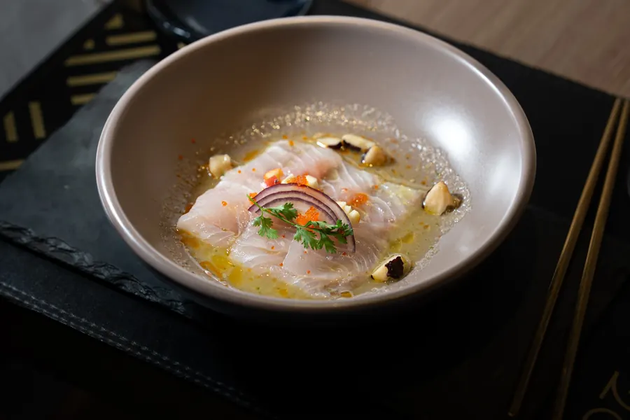 Lâminas de peixe branco cru em caldo cítrico, com cebola roxa, coentro, ovas e castanhas, numa tigela cinza sobre ardósia, com hashis dourados ao lado.
